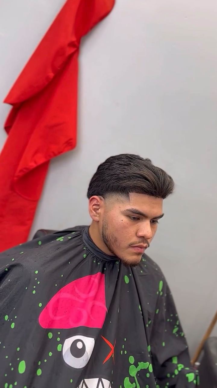 Client with a low taper fade and longer dark hair styled up and back, with a trimmed beard